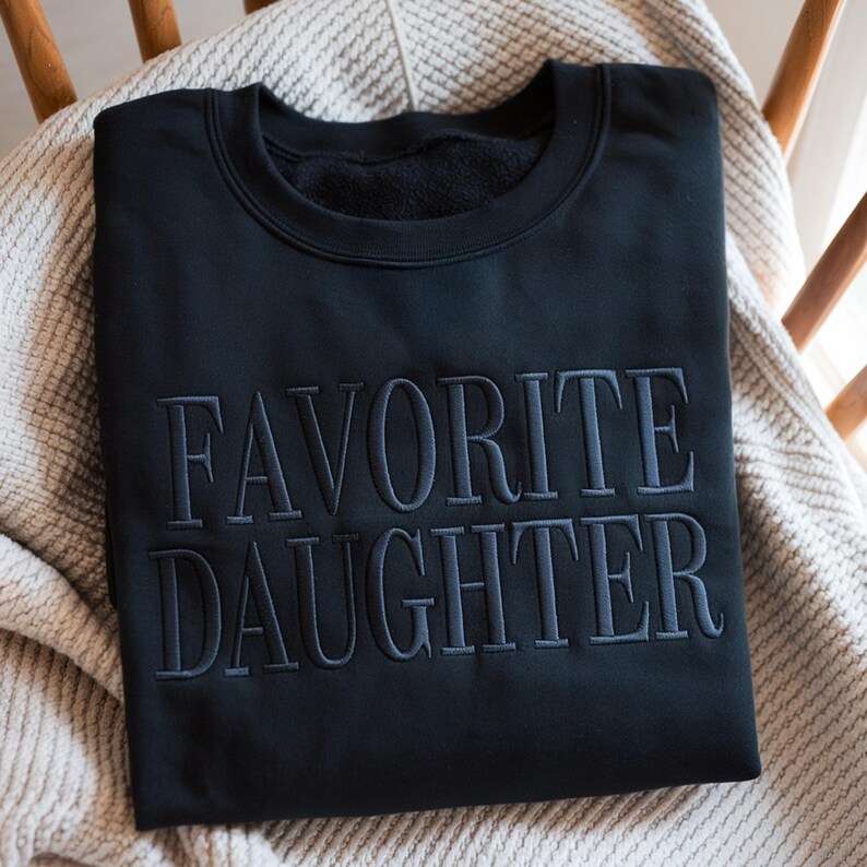 May include: Black sweatshirt with the embroidered text "FAVORITE DAUGHTER" in a darker shade of gray. The sweatshirt is folded and resting on a light-colored blanket, placed on a wooden chair.