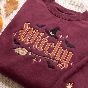 May include: A maroon crewneck sweatshirt featuring embroidered text that reads "witchy" in a stylized font. The design includes a black witch hat, a brown broomstick, several small black bats, and gold-colored stars scattered around the lettering.