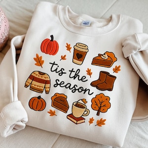 May include: Cream-colored sweatshirt with embroidered fall-themed graphics. The design includes a pumpkin, sweater, coffee cup, pie, boots, and leaves. The phrase "tis the season" is written in black script.