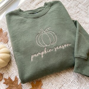 May include: An olive green crewneck sweatshirt featuring a white embroidered line art illustration of a pumpkin centered on the chest, with the text "pumpkin season" embroidered in a cursive white font directly below the pumpkin graphic.