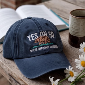 May include: A navy blue baseball cap with the text "YES ON 50" and a brown bear graphic. The phrase "DEFEND DEMOCRACY CALIFORNIA" is embroidered below the bear. The cap is sitting on a wooden surface.