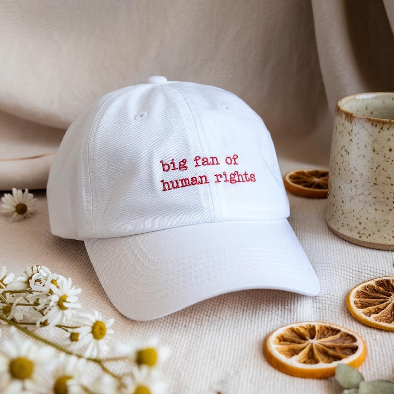 A white dad hat embroidered in red lowercase letters with the phrase “big fan of human rights,” styled on a neutral linen backdrop with dried citrus, chamomile flowers, and a ceramic mug.