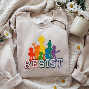 Cream sweatshirt with colorful rainbow character silhouettes embroidered above bold “RESIST” text. Displayed with daisies, glasses, and a mug on a soft neutral knit blanket.