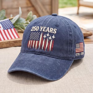 May include: A navy blue baseball cap with the text "250 YEARS" above a distressed American flag design with jets. The cap also features a small American flag patch with the dates 1776-2026.