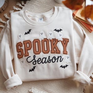 May include: Cream-colored sweatshirt with the words "SPOOKY Season" in brown and black lettering. The sweatshirt features embroidered white ghosts and black bats, perfect for Halloween.