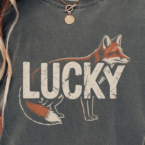 Puede incluir: Camiseta unisex color pimienta con un gráfico "LUCKY" desgastado y una ilustración de zorro en tonos marrón y blanco. La camiseta tiene mangas cortas y cuello redondo.