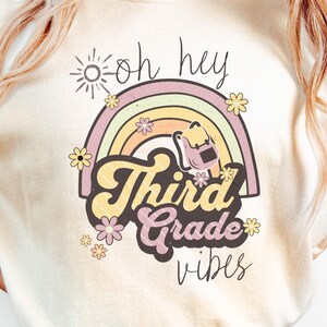 Op de afbeelding: Een ivoorkleurig t-shirt met een grafische print van een regenboog, een kleine rugzak, bloemen, een zon en de tekst "oh hey Third Grade vibes". Het ontwerp bevat zachte pastelkleuren zoals geel, roze en lichtblauw op de lichte stof.