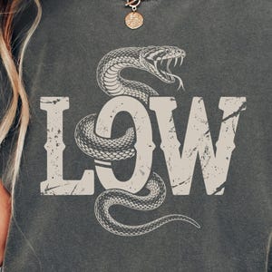Puede incluir: Camiseta unisex color pimienta con un gráfico desgastado de una serpiente enrollada alrededor de la palabra "LOW". El diseño es de estilo vintage. La camiseta es cómoda y de uso casual.