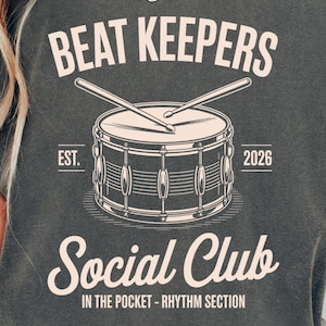 Può includere: T-shirt unisex color pepe con la scritta "BEAT KEEPERS Social Club" e una grafica di batteria. Il design include il testo "EST. 2026" e "IN THE POCKET - RHYTHM SECTION". La maglietta è uno stile comodo e casual.