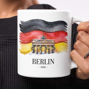 May include: White ceramic mug featuring a watercolor illustration of the Brandenburg Gate in Berlin, Germany, with the German flag in the background. The word "BERLIN" is printed below the image, along with the year "- 2026 -."