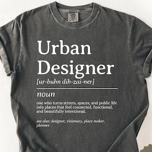Może przedstawiać: Ciemnoszara koszulka z białym napisem "Urban Designer", przewodnikiem po wymowie i definicją. Koszulka zawiera również słowo "noun" i powiązane terminy. Koszulka jest oznaczona jako "Pepper".