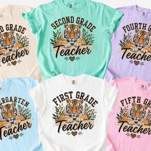 Puede incluir: Seis camisetas de manga corta en colores marfil, arrecife, orquídea, chambray, blanco y flor. Cada camiseta presenta un gráfico de un tigre descansando sobre una rama con hojas, texto que indica el nivel escolar desde jardín hasta quinto grado y un corazón.