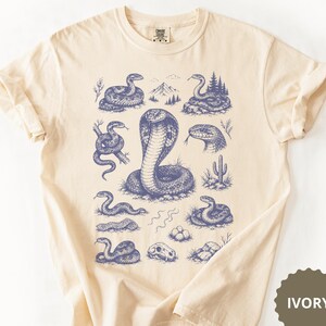 Peut inclure: Un t-shirt ivoire avec une impression graphique bleue représentant divers serpents, dont un cobra central, un crâne de serpent, un cactus et un paysage de montagne. Le motif est disposé en grille sur le devant du t-shirt, montrant des illustrations détaillées de reptiles dans différentes positions.
