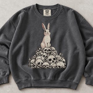 May include: A charcoal gray crewneck sweatshirt featuring a graphic print of a light-colored rabbit sitting atop a large pile of human skulls and bones. The garment has long sleeves, a ribbed collar, and a Comfort Colors brand label at the neckline.