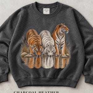 Pode incluir: Uma camisola de gola redonda em cinzento antracite mesclado com uma impressão gráfica de três tigres, incluindo um tigre branco, a beber de um corpo de água com erva alta e rochas. A etiqueta interior exibe o texto Lane Seven e tamanho L.