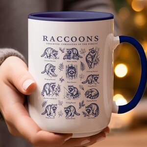 White mug with navy blue rim and handle featuring diverse raccoon illustrations and text.