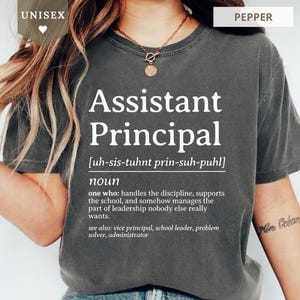 May include: A charcoal gray short-sleeve t-shirt featuring white text that defines Assistant Principal as a noun, including a phonetic pronunciation and a humorous description of the role, with the word UNISEX and PEPPER labels visible.