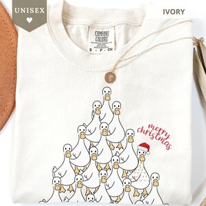 Może przedstawiać: Kremowa koszulka unisex z nadrukiem choinki z kaczuszek z kreskówek. Kaczki są białe z żółtymi dziobami i stopami. Jedna kaczka ma na sobie czapkę Świętego Mikołaja. Napis "merry christmas" jest w kolorze czerwonym.