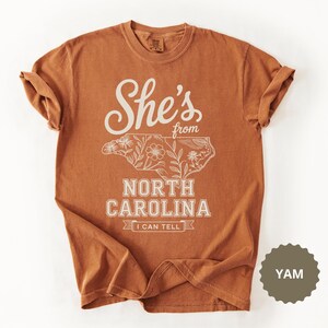 Brown t-shirt with "She's from North Carolina" text and floral outline of the state.