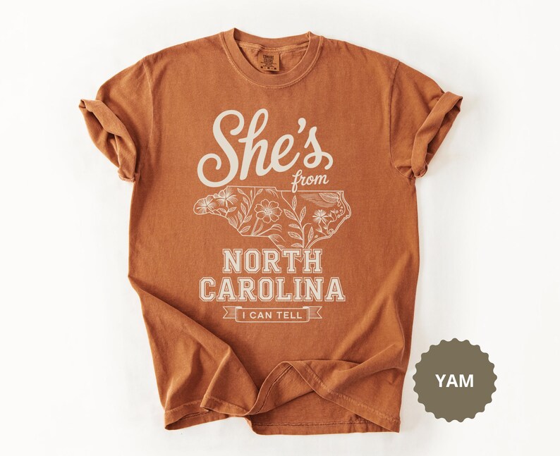 Brown t-shirt with "She's from North Carolina" text and floral outline of the state.