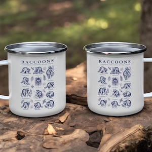 Two white enamel mugs with blue raccoon illustrations and text.