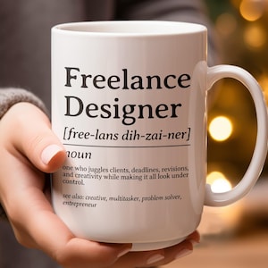 May include: A white ceramic mug with the words "Freelance Designer" printed in black. The mug includes a definition and related terms. The mug is held in a person's hand.