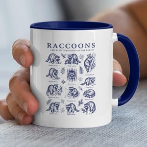 White mug with blue trim featuring a collage of raccoon illustrations and labels.