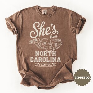 Brown t-shirt with "She's from North Carolina I Can Tell" text and floral outline map.