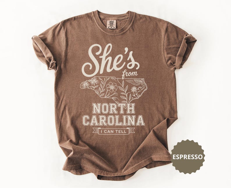 Brown t-shirt with "She's from North Carolina I Can Tell" text and floral outline map.
