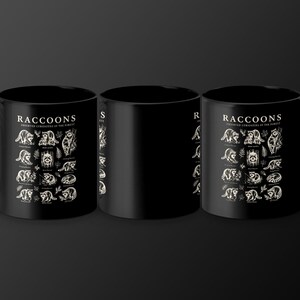 Black mugs with a detailed raccoon illustration and text "Raccoons Observed Curiosities of the Forest".