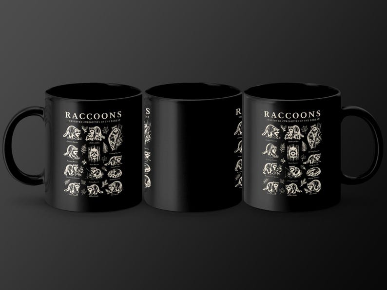 Black mugs with a detailed raccoon illustration and text "Raccoons Observed Curiosities of the Forest".