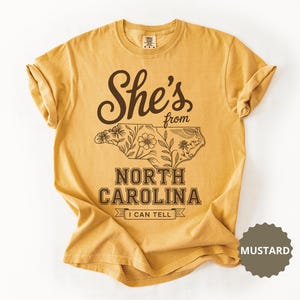 Mustard t-shirt with floral outline of North Carolina and "She's from North Carolina I can tell" text.
