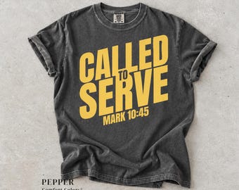 Può includere: Una t-shirt a maniche corte color grigio antracite con scollo rotondo. La parte anteriore presenta una scritta gialla in grassetto che recita CALLED TO SERVE e MARK 10:45. Il tessuto ha un aspetto slavato e vintage con maniche arrotolate.