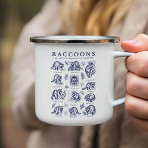White enamel mug with dark blue illustrations and text about raccoons observed in the forest.