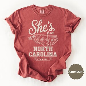 Crimson t-shirt with "She's from North Carolina" and floral outline of the state.