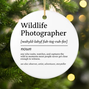 May include: A white, round Christmas ornament with the text "Wildlife Photographer" in black. The definition of the term is also included, along with related terms. A gold string is attached for hanging.