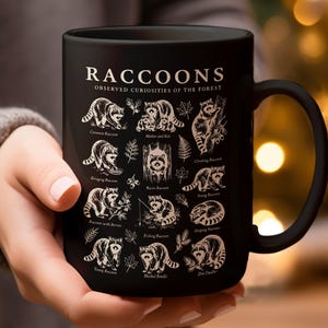 Black mug with raccoon illustrations and "Raccoons Observed Curiosities of the Forest" text.