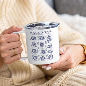 White mug with dark blue print of raccoons and text "Raccoons Observed Curiosities of the Forest."