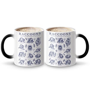 Two white mugs with blue raccoon illustrations and the text "Raccoons Observed Curiosities of the Forest."