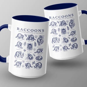 Two white mugs with navy blue rims and handles, featuring detailed raccoon illustrations and text.