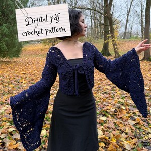 Può includere: Un bolero all'uncinetto blu navy con maniche a campana. Il bolero è legato sul davanti e presenta un delicato design traforato. L'immagine include un foglio con il testo "Digital pdf crochet pattern". Lo sfondo presenta foglie autunnali e alberi.