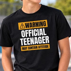 May include: A black short-sleeved t-shirt featuring a yellow and white graphic print that reads WARNING OFFICIAL TEENAGER MAY CONTAIN ATTITUDE with a warning triangle icon. The design is centred on the chest of the garment.