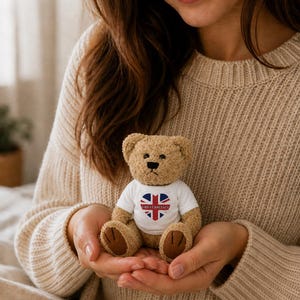 May include: A small, light brown plush teddy bear with dark brown paw pads. The bear wears a white t-shirt featuring a heart-shaped Union Jack flag design with the text GREAT BRITAIN printed in black letters across the centre of the heart.