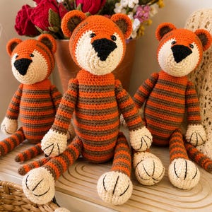 May include: Three handmade crochet tiger toys featuring orange and brown horizontal stripes, cream-coloured faces, black triangular noses, and dark-tipped ears. Each stuffed animal has long limbs and cream-coloured paws with stitched details on the feet.