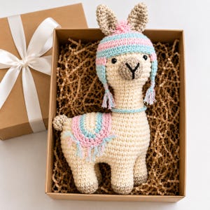 Può includere: Un giocattolo a forma di lama lavorato all'uncinetto color crema, che indossa un cappello a righe azzurre e rosa con nappe e una coperta da sella abbinata con frange. Il lama è inserito in una scatola regalo in cartone marrone con paglia.