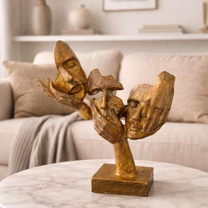 Könnte beinhalten: Eine goldfarbene Skulptur mit drei stilisierten Gesichtern, die von einer Hand gehalten werden. Die Skulptur ist auf einem quadratischen Sockel montiert und steht auf einem Tisch mit Marmorplatte. Der Hintergrund umfasst ein Sofa und dekorative Elemente.