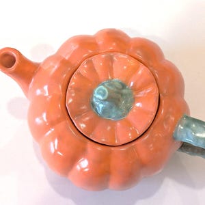 May include: A ceramic teapot shaped like a pumpkin, with a glossy orange finish. The lid and handle are a muted teal colour. The teapot has a spout and a handle, and is designed for brewing tea.