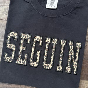 以下が含まれることがあります： 胸元に大きなブロック体で「SEGUIN」という文字がプリントされた黒の半袖Tシャツ。文字はベージュと茶色のヒョウ柄で、太い黒の縁取りが施されている。首元の内側のラベルには「COMFORT COLORS L G G」と記されている。