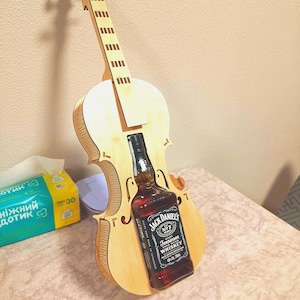 May include: A wooden violin-shaped bottle holder with a bottle of Jack Daniel's Tennessee Whiskey. The holder is made of light-colored wood and has a cutout to accommodate the bottle. The whiskey bottle is dark with a black and white label.