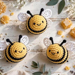 May include: Three round, crocheted bee figures featuring yellow and black horizontal stripes, small black antennae, and delicate white lace-like wings. Each bee has a simple stitched smile and two small black eyes, arranged on a white surface.